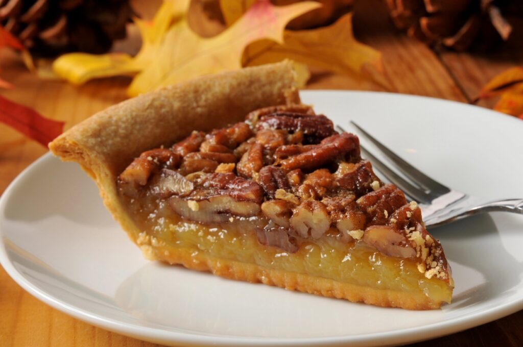 A slice of pecan pie on a white plate with a fork, set on a wooden table. The pie has a golden crust and is topped with whole pecans. Autumn leaves and pinecones are in the background, creating a cozy, fall atmosphere.