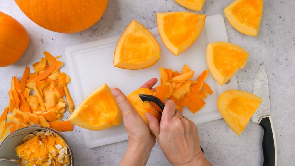 Person peeling slices of pumpkin on a white cutting board. There are seeds and peel in a bowl to the side, along with a knife and additional pumpkin pieces on the table.