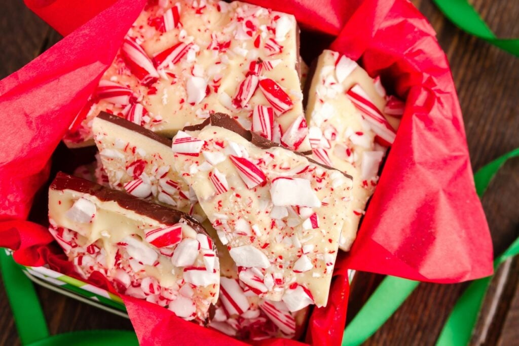 A festive gift box filled with pieces of peppermint bark wrapped in red tissue paper. The bark features a layer of white chocolate topped with crushed candy canes and a layer of dark chocolate underneath, set against a wooden background with green ribbon accents.