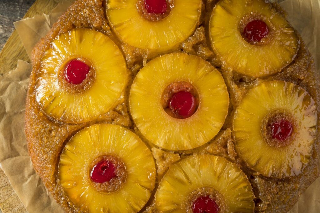 A close-up of a pineapple upside-down cake topped with slices of pineapple and maraschino cherries. The cake has a golden-brown surface and is placed on parchment paper.