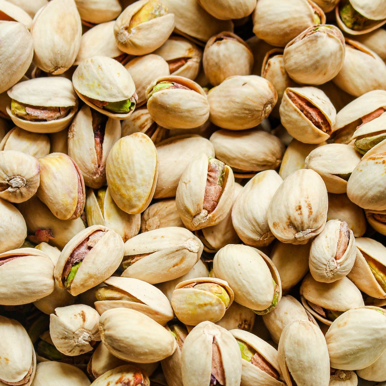 Close-up of a pile of unshelled pistachios, showcasing their beige shells with green and purple-tinted nuts peeking through the openings. The image highlights the texture and natural colors of the pistachios.