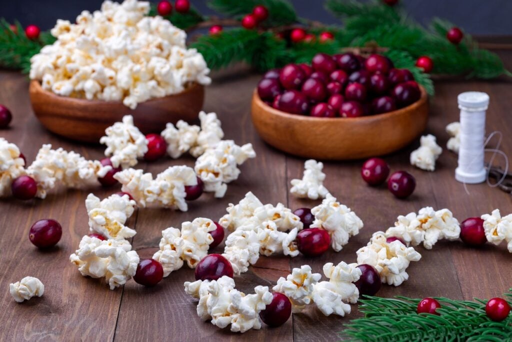 Bowls of popcorn and cranberries are scattered on a wooden table. Sprigs of evergreen and a spool of white thread add a festive touch, with popcorn and cranberries arranged in a garland around the scene.