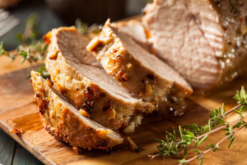 Slices of roasted pork with a golden-brown crust on a wooden cutting board, garnished with fresh thyme sprigs.