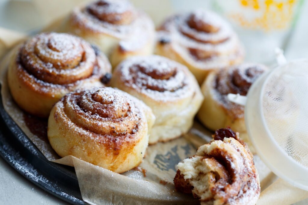 A tray of freshly baked cinnamon rolls dusted with powdered sugar. One roll is partially torn, showing its soft, swirled interior. They rest on parchment paper with a sugar sifter nearby.