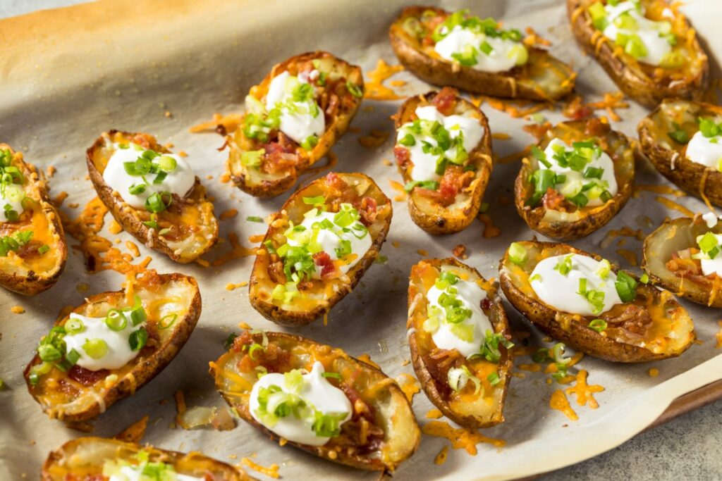 Baked potato skins topped with melted cheese, crispy bacon, sour cream, and chopped green onions on a parchment-lined baking sheet.