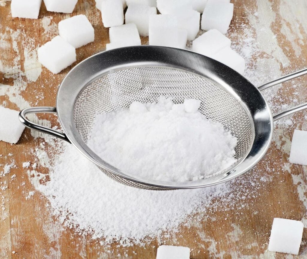 A metal sifter containing powdered sugar sits on a wooden surface, surrounded by scattered sugar cubes and more powdered sugar. The scene suggests baking preparation.