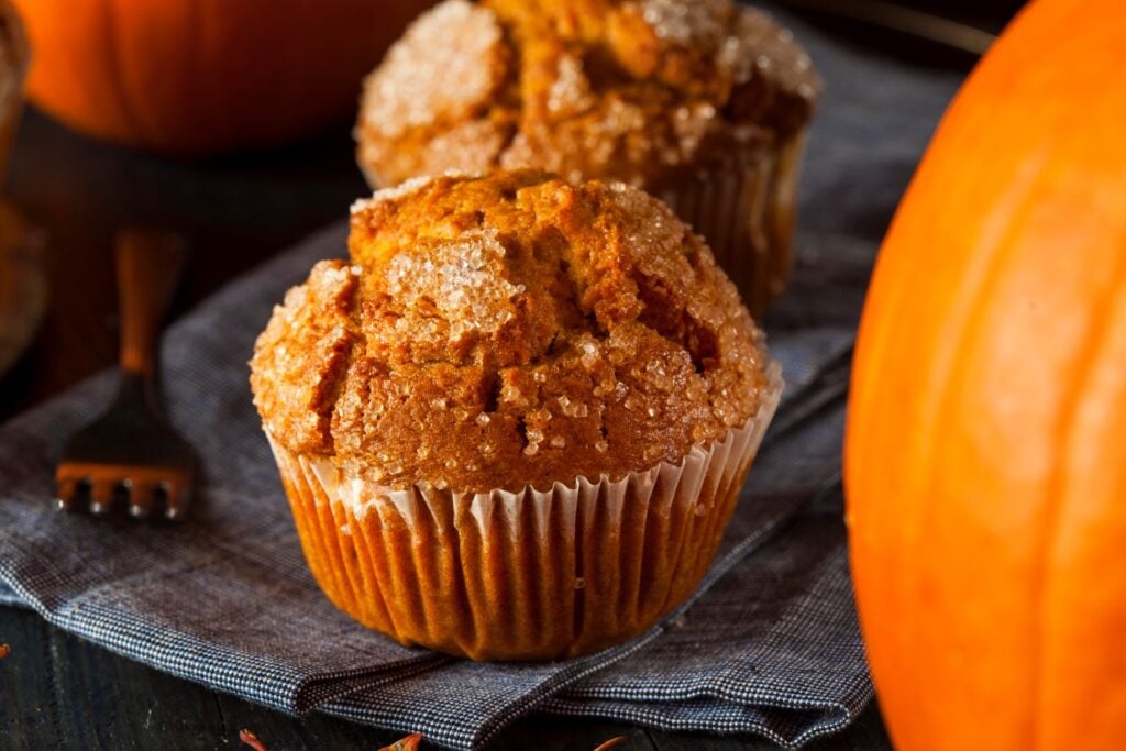 Close-up of two pumpkin muffins with sugar crystals on top, sitting on a blue cloth. A whole pumpkin is partially visible on the right. A fork rests nearby, highlighting a cozy autumn setting.