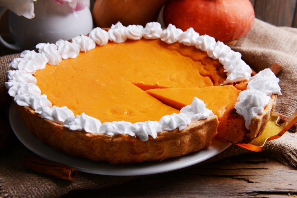 A pumpkin pie with a missing slice sits on a white plate. The pie is topped with whipped cream around the crust, and a golden server holds the cut slice. In the background, there are scattered autumn decorations, including a pumpkin.