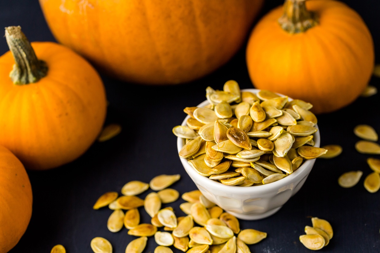 A small white bowl filled with roasted pumpkin seeds sits on a dark surface, surrounded by scattered seeds. In the background, three whole pumpkins add a vibrant orange contrast.