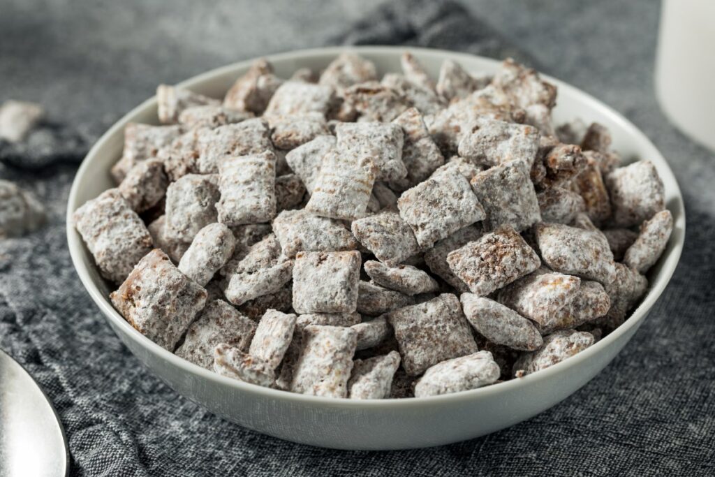 A bowl filled with cereal squares coated in powdered sugar. The cereal appears to be a sweetened snack mix, and it rests on a dark cloth surface.