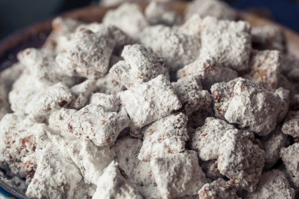 A close-up of a pile of powdered sugar-coated snacks, resembling small bite-sized squares. The texture appears crunchy and the sugar adds a white, snowy coating over each piece, giving a sweet and enticing appearance.