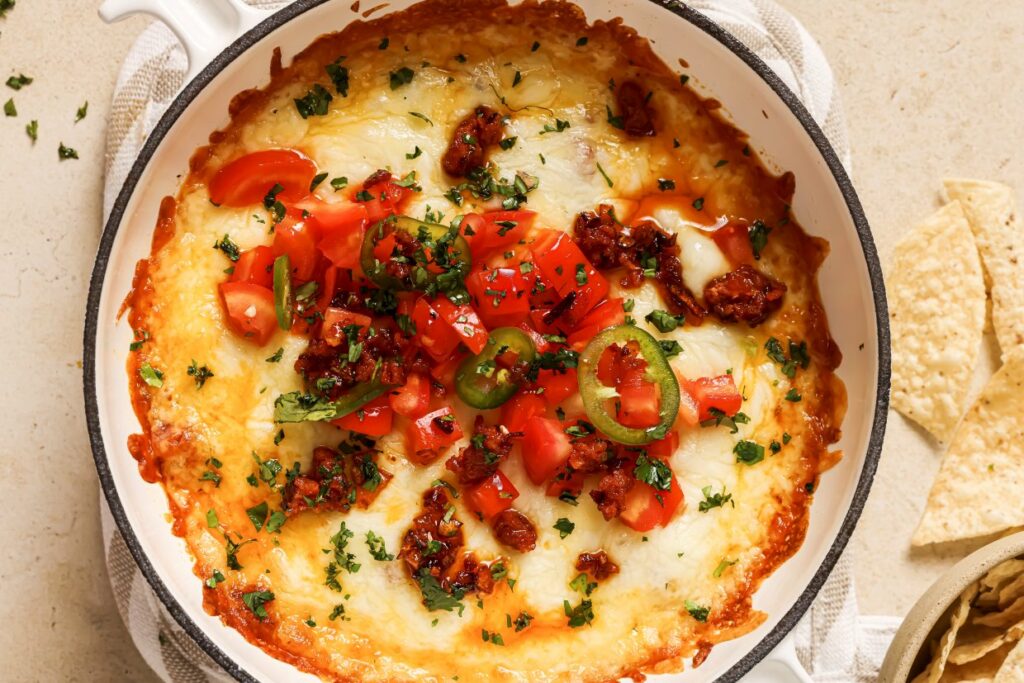 A baked dish topped with melted cheese, diced tomatoes, jalapeño slices, and crumbled chorizo, garnished with chopped cilantro. The dish is in a round, white baking dish surrounded by tortilla chips.