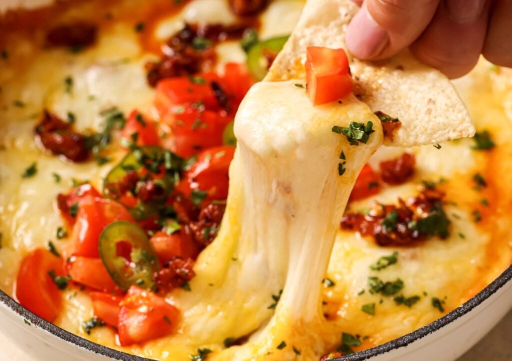 A close-up of a cheesy dip topped with diced tomatoes, jalapeños, and herbs. A hand is dipping a tortilla chip into the melted cheese, stretching it out. The dish appears appetizing and colorful.