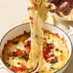 A hand scoops a tortilla chip covered in gooey cheese from a casserole dish with queso fundido.