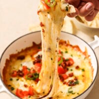 A hand scoops a tortilla chip covered in gooey cheese from a casserole dish with queso fundido.