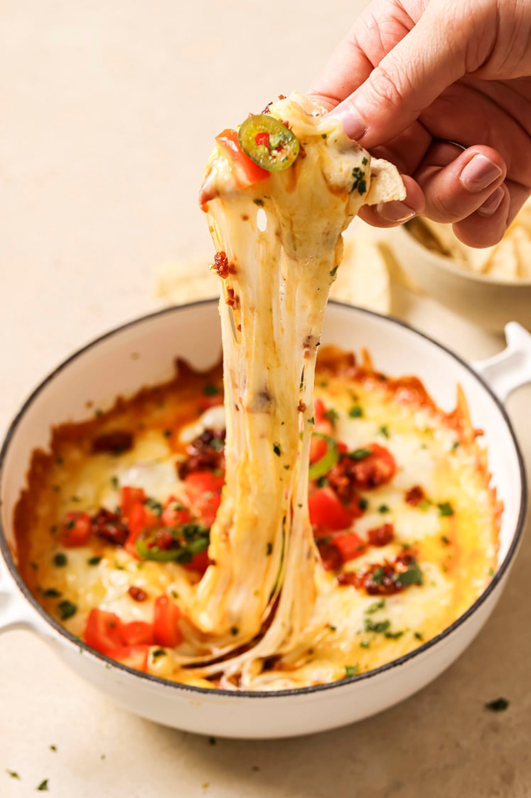 A hand scoops a tortilla chip covered in gooey cheese from a casserole dish with queso fundido.