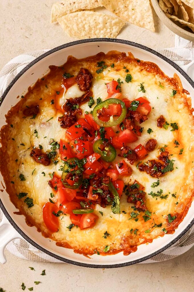 A close-up of a queso fundido topped with diced tomatoes, jalapeño slices, cilantro, and pork chorizo on a small casserole dish.