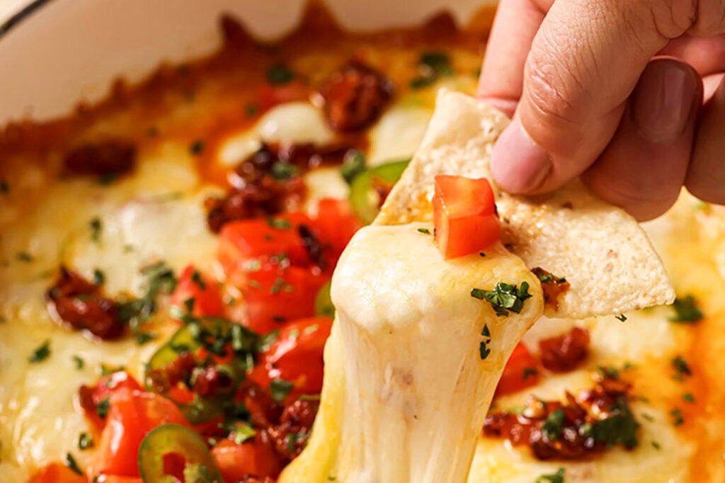 A hand scoops a tortilla chip covered in gooey cheese from a casserole dish with queso fundido.