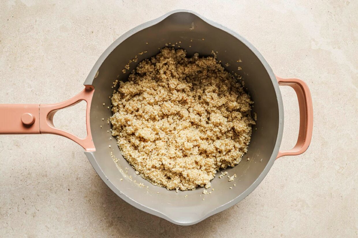 A pot filled with cooked quinoa sits on a light-colored surface. The quinoa is fluffy and evenly spread, and the pot has a pink handle.