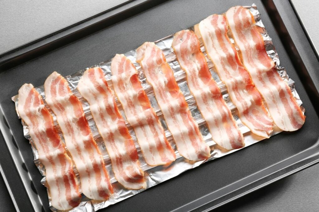 Raw bacon strips are neatly arranged on a foil-lined baking sheet, ready for cooking.