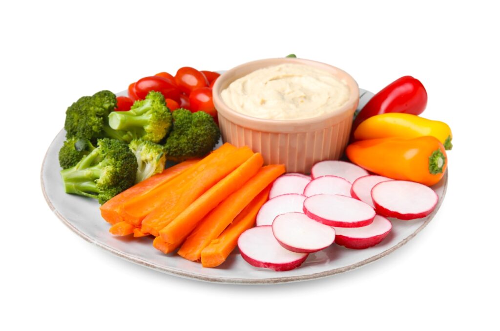 A plate of fresh vegetables including broccoli, carrot sticks, red radish slices, cherry tomatoes, and mini bell peppers arranged around a bowl of creamy dip. The plate is set against a white background.