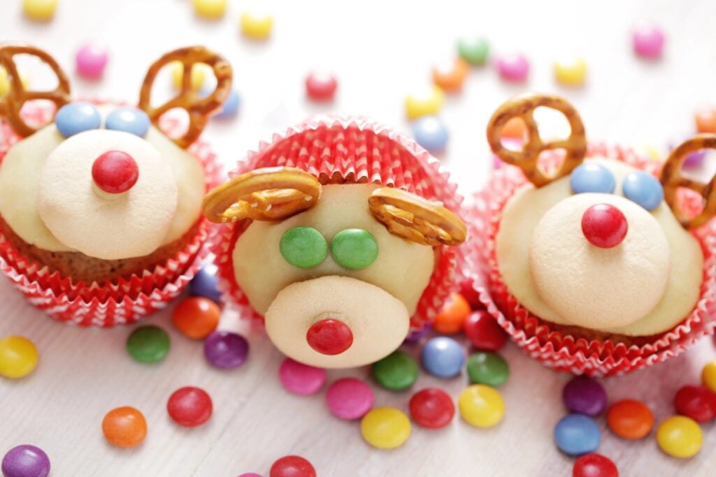 Three cupcakes decorated to look like reindeer. Each has a pretzel for antlers, colored candy for eyes and nose, and is set in a red cupcake liner. Colorful candies are scattered around them on a white surface.