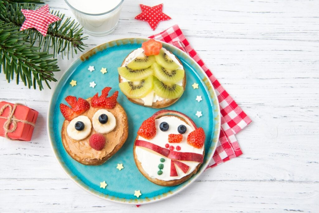 A festive plate with three decorated cookies: a reindeer with blueberries, a kiwi Christmas tree, and a snowman with strawberry accents. Nearby are a glass of milk, a small gift, and pine branches, all on a white wooden table.