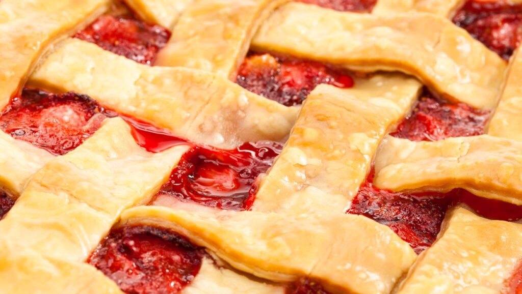 Close-up of a lattice-topped rhubarb pie, showcasing golden-brown, crisscrossed pastry strips over a filling of vibrant red strawberries and syrup. The texture is glossy and slightly bubbly, suggesting it was freshly baked.