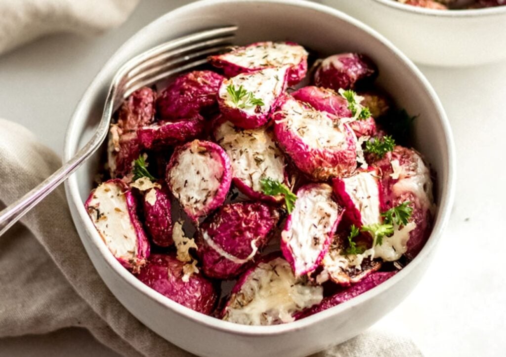 A bowl of roasted radishes topped with melted cheese and garnished with parsley. A fork rests on the edge of the bowl, and a beige napkin is partially visible beside it.