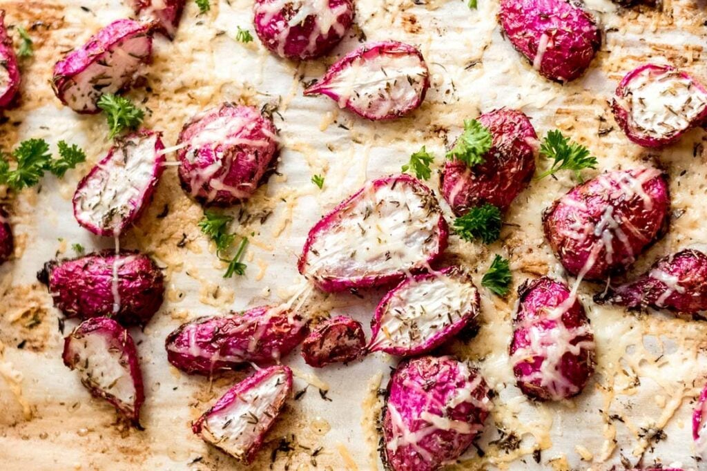 Roasted radishes topped with shredded cheese and herbs on a baking sheet, with sprigs of fresh parsley scattered among them.