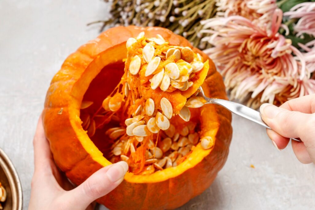 A person scoops out seeds and pulp from a halved pumpkin with a spoon. The pumpkin rests on a table, surrounded by decorative flowers. The orange interior and seeds are clearly visible.