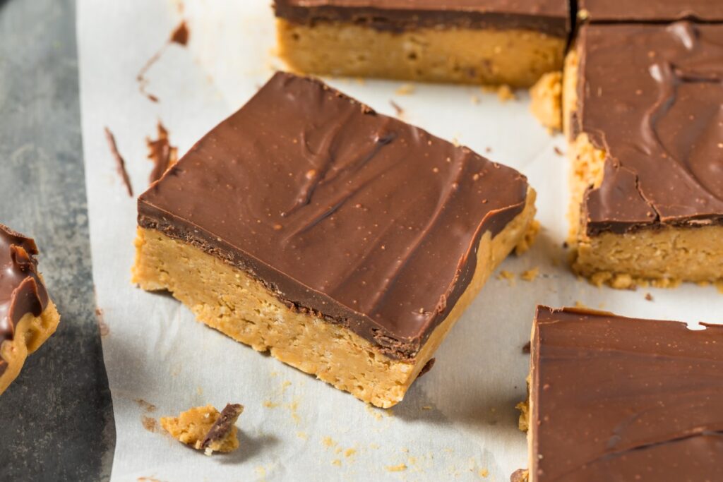 A close-up of chocolate peanut butter bars on parchment paper. The bars have a layered appearance with a thick peanut butter base and a smooth chocolate topping, cut into square pieces.