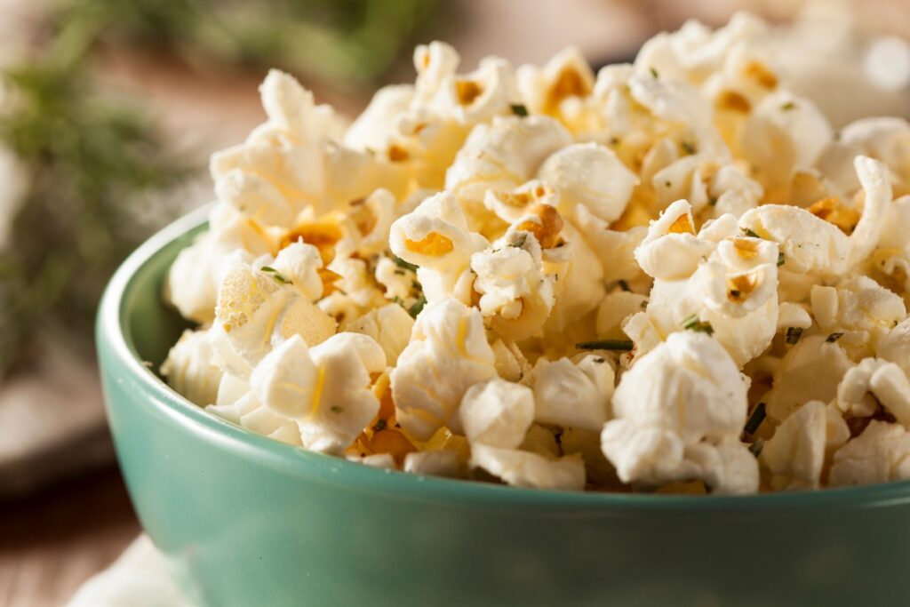 A green bowl filled with freshly popped popcorn, garnished with herbs. The popcorn is fluffy and lightly golden, creating an appetizing look. The background is blurred, emphasizing the bowl of popcorn in the foreground.