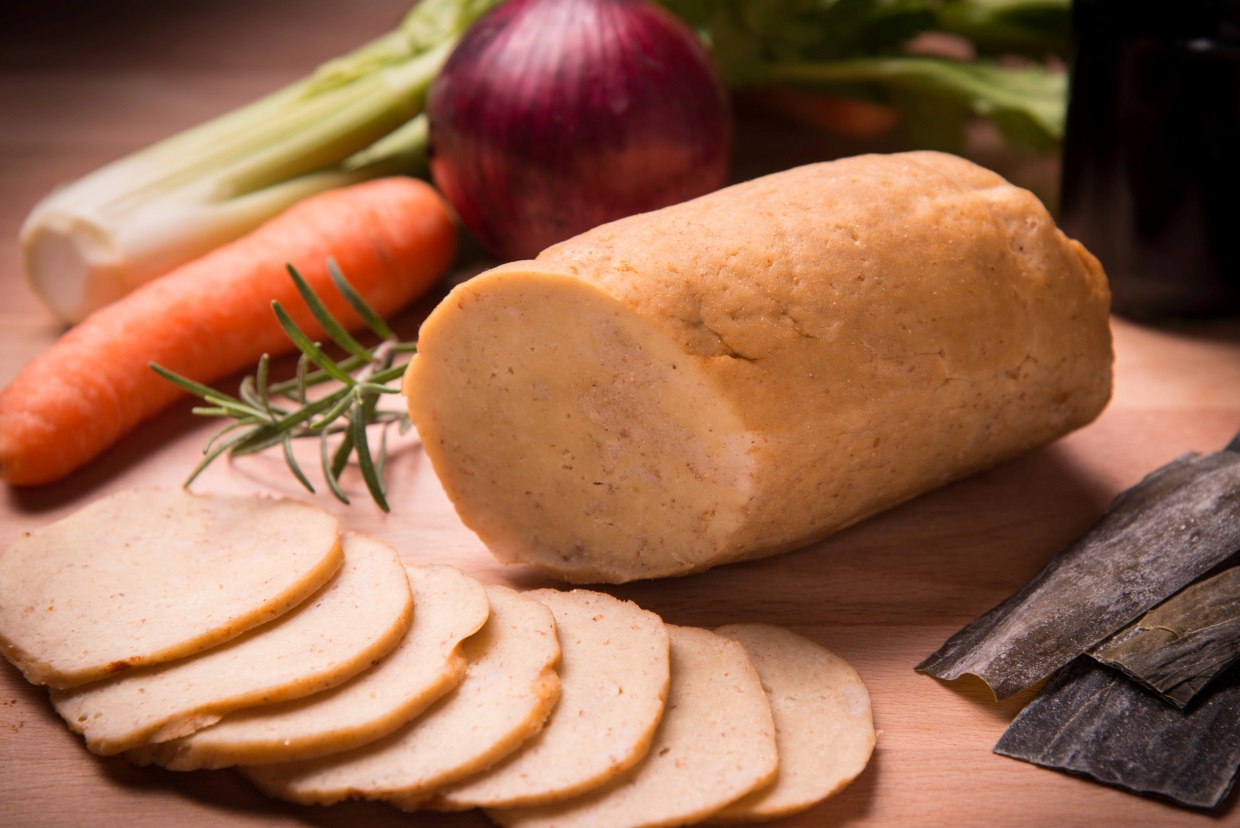 A round loaf of sliced seitan is displayed on a wooden surface with rosemary, a carrot, a red onion, celery stalks, and dried seaweed pieces in the background.