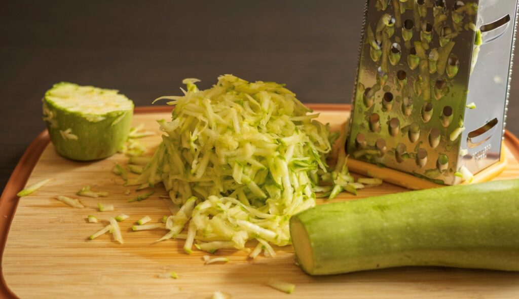 Grated zucchini piled on a wooden cutting board next to a metal grater. A partially sliced zucchini and a zucchini end are also on the board.