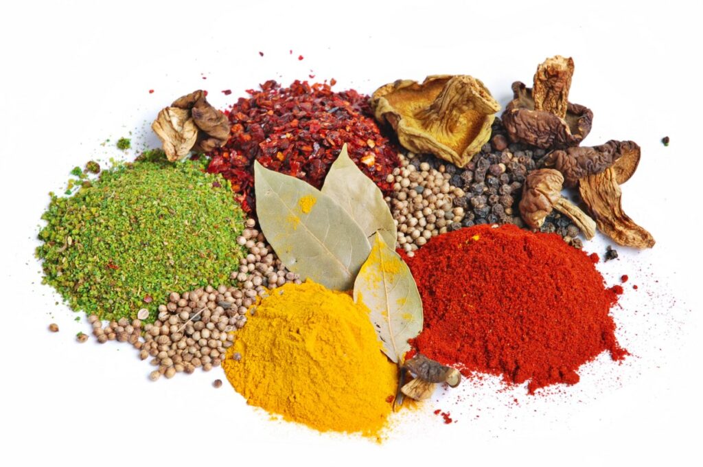 Assorted spices arranged on a white background, including green herb mix, red chili flakes, dried mushrooms, black peppercorns, turmeric powder, coriander seeds, paprika, and bay leaves.