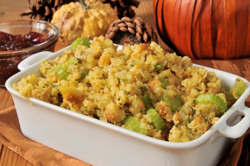 A white casserole dish filled with golden brown stuffing with visible celery pieces, surrounded by autumn-themed decor, including pine cones, a small pumpkin, and a dish of cranberry sauce.