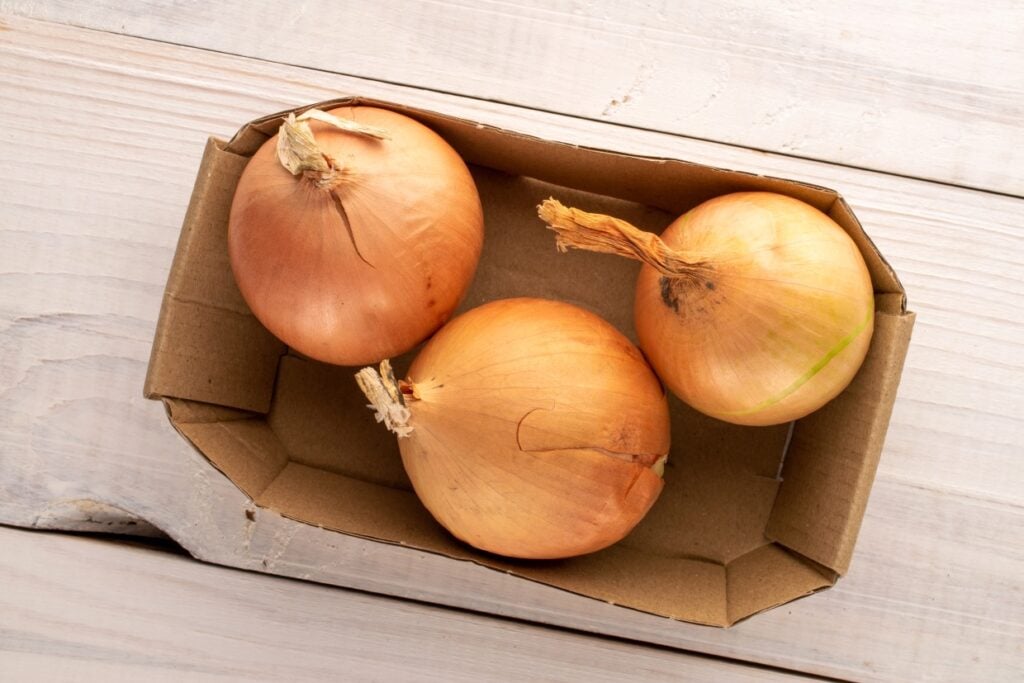 Three brown onions in a cardboard container on a light wooden surface, viewed from above.