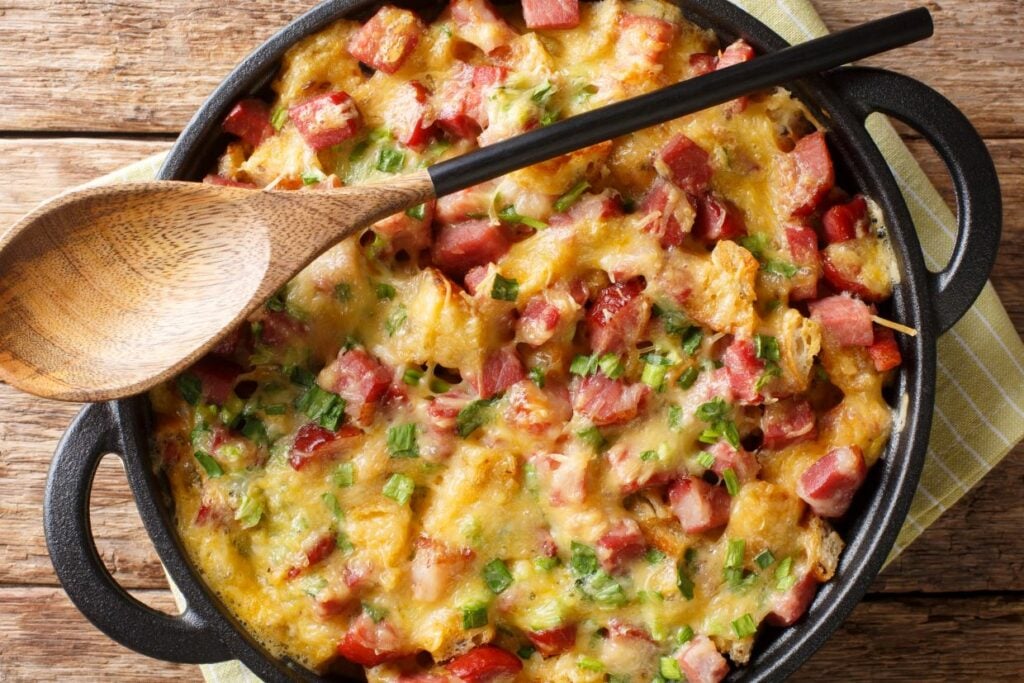 A baked casserole in a black dish on a wooden surface, topped with melted cheese, diced ham, and green onions. A wooden spoon rests on the dish's edge.