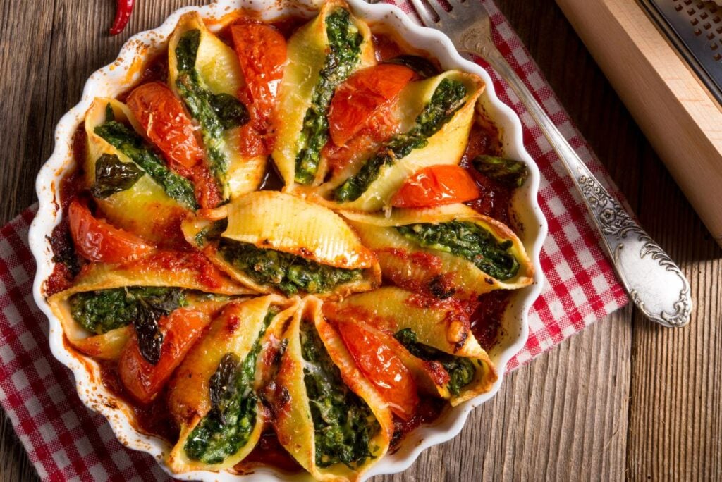 A round baking dish filled with stuffed pasta shells, containing spinach and tomatoes, baked with a cheese topping. The dish is on a checkered cloth, with a decorative spoon and a grater nearby, placed on a wooden surface.