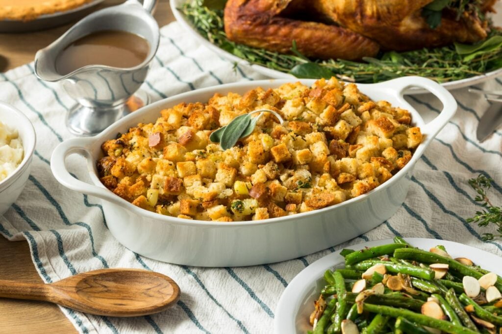 A white casserole dish filled with golden-brown stuffing garnished with herbs. It sits on a striped cloth alongside a small gravy boat and a serving of green beans with almonds. A roasted turkey is partially visible in the background.