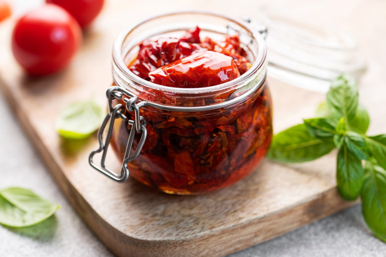 A glass jar filled with sun-dried tomatoes in oil sits on a wooden board, surrounded by fresh basil leaves and partially visible cherry tomatoes. The lid rests nearby, showcasing the vibrant colors and textures of the ingredients.