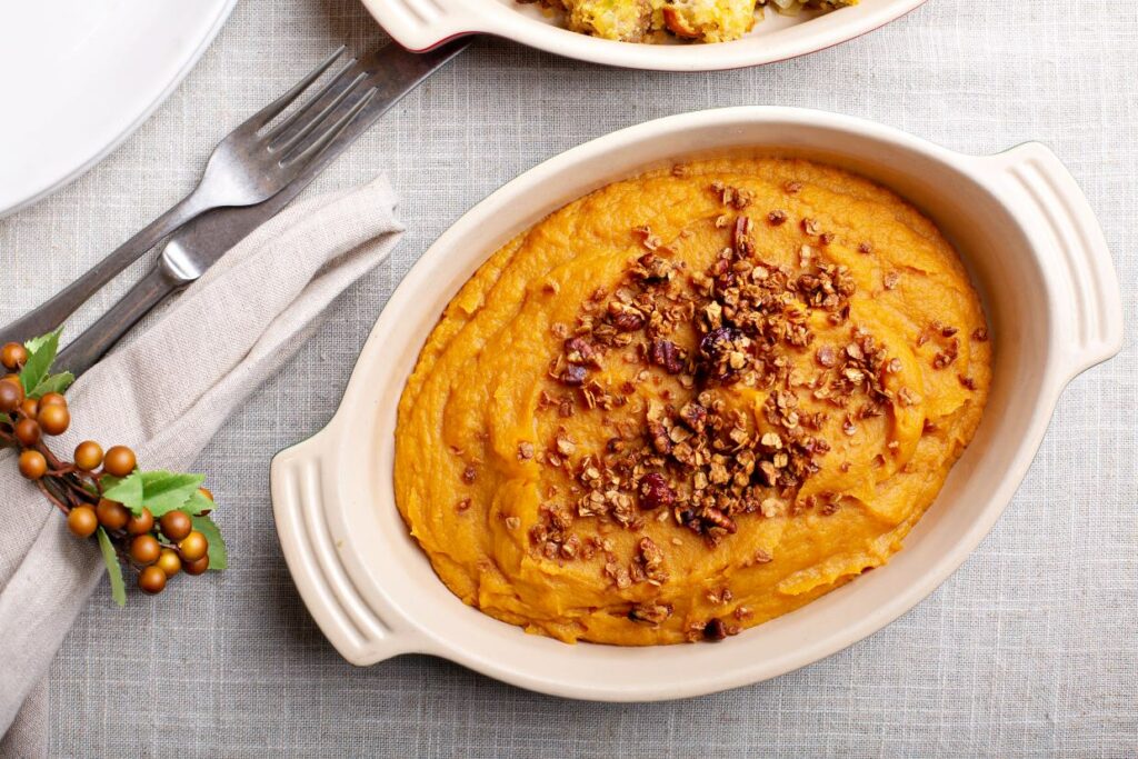 An oval dish with creamy sweet potato casserole topped with crushed pecans, set on a light gray woven tablecloth. A fork and knife rest on a beige napkin beside the dish, with a small sprig of berries nearby.