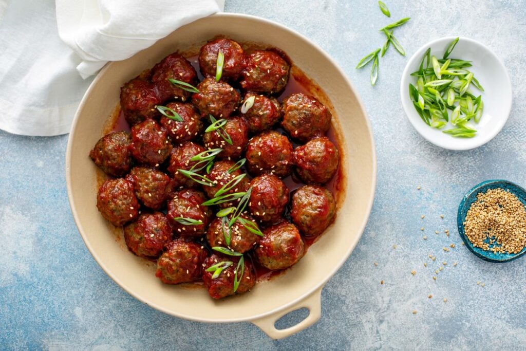 A round dish filled with glazed meatballs, garnished with sliced green onions and sesame seeds. Nearby are bowls of chopped green onions and sesame seeds. The background is a textured blue surface, partially covered with a white cloth.