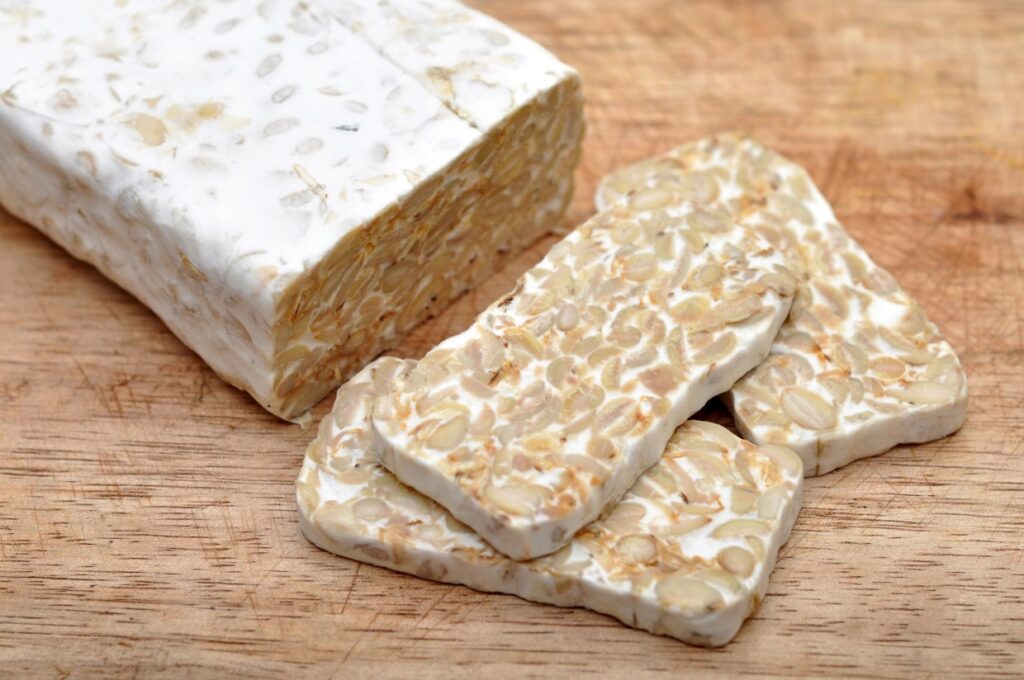 A block of tempeh is partially sliced on a wooden cutting board. The tempeh has a textured appearance, with visible soybeans bound together by a white, fuzzy coating. Three slices are laid out next to the main block.