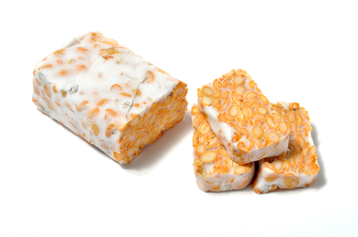 A block of tempeh is shown with three slices next to it on a white background. The tempeh has a firm texture and the soybeans are visible within it.