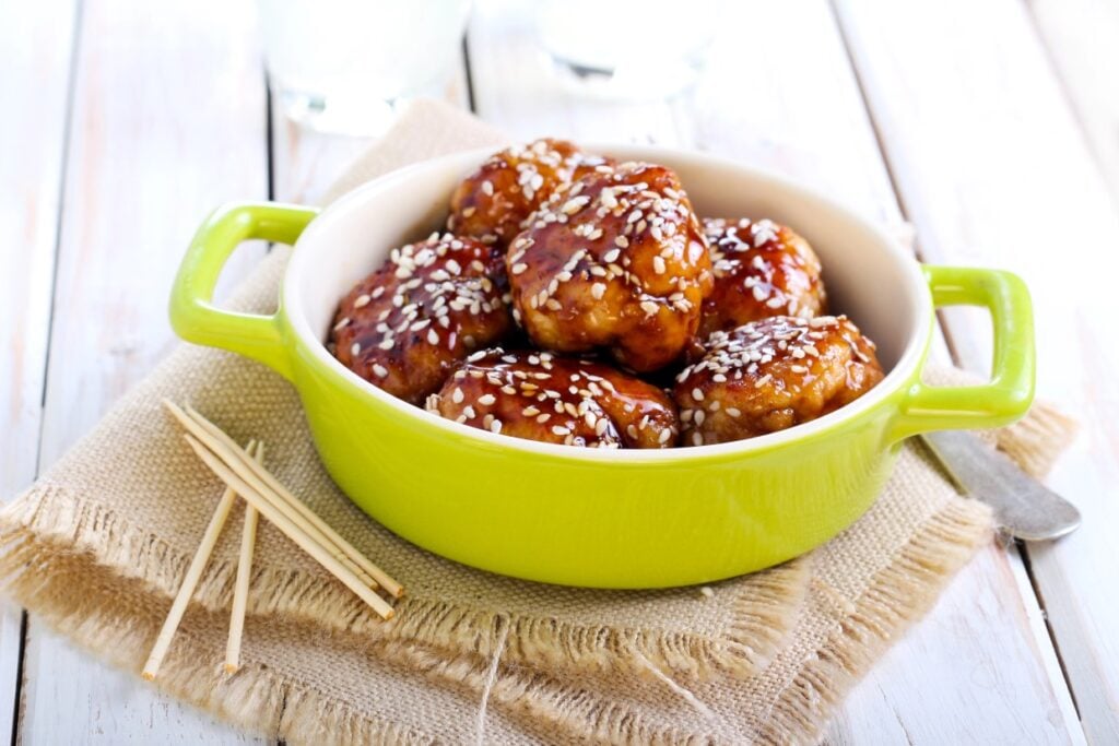 A green bowl filled with glazed meatballs topped with sesame seeds sits on a burlap cloth. Two wooden skewers lie beside the bowl on a white wooden table.