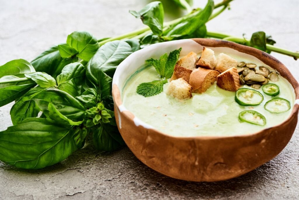 A bowl of green soup garnished with croutons, pumpkin seeds, mint, and sliced jalapeños, served with fresh spinach leaves on the side. The soup is in a rustic brown bowl on a textured surface.