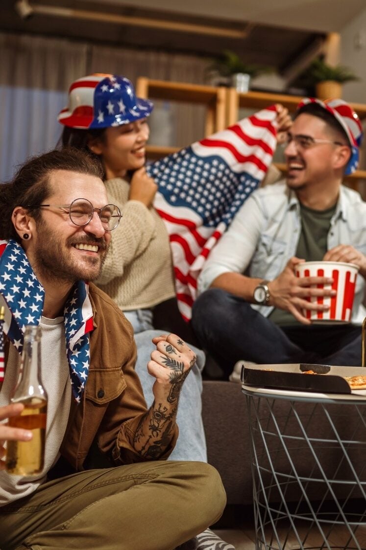 Three people celebrate indoors, wearing flag-themed hats and scarves. One holds popcorn, another a bottle, and they appear happy and engaged. The setting is cozy with modern decor, and they are likely watching a sporting event.