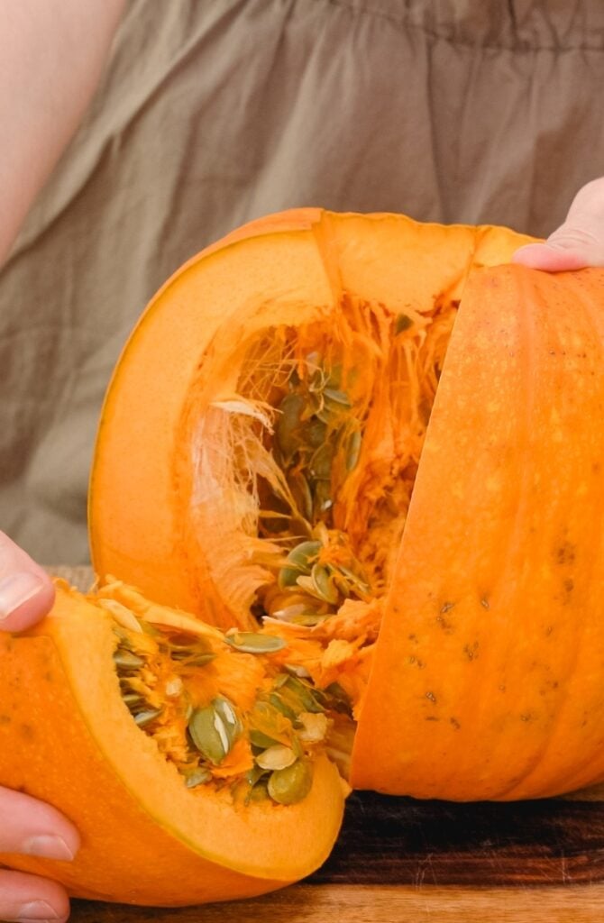 A person is holding a halved pumpkin, revealing its seeds and stringy insides. The pumpkin is bright orange, and the person is wearing a brown shirt.